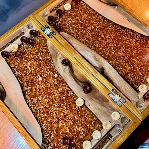 May include: A wooden backgammon board with a warm, amber-colored playing surface. The board features brown and tan game pieces, with a wooden frame. The game board is set on a wooden table, with a drink in the background.