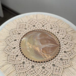 Puede incluir: Un mantel individual redondo de crochet de color crema con un borde de flecos. El mantel individual tiene un diseño decorativo intrincado y se coloca sobre una superficie blanca.