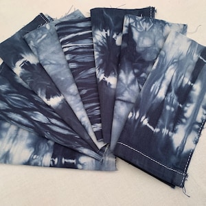 May include: A selection of indigo blue tie-dye fabric napkins, arranged to show the different patterns. The napkins feature white stitching on the edges and a range of swirling and striped designs.