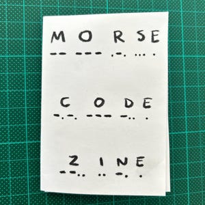 May include: A white card with the words "MORSE CODE ZINE" written in black marker. Below each word is a series of dashes and dots representing the Morse code for each letter.
