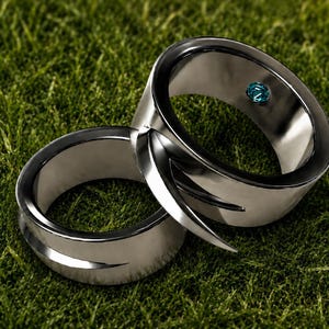 May include: Two polished silver-toned metal rings with a smooth, reflective finish. The rings feature a distinctive overlapping design with pointed, curved edges. One ring is accented with a single, small, round, bright blue gemstone.