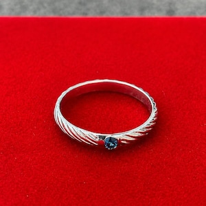 May include: A silver ring featuring a twisted cable texture along the band and a single, small, round, deep blue gemstone set at the front. The metal has a polished, reflective finish that catches the light, highlighting the intricate grooves.