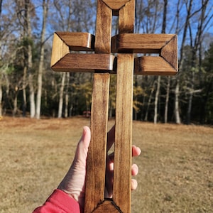 May include: A handcrafted wooden cross with a rich, dark brown stain. The cross features a unique, layered design with geometric shapes and a smooth finish. The cross is held against a backdrop of trees and a clear blue sky.