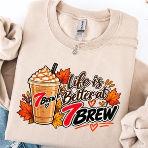 May include: A beige crewneck sweatshirt featuring a graphic of an iced coffee drink with whipped cream and caramel drizzle. The text reads "Life is Better at 7 BREW" alongside orange and red autumn maple leaves and small orange heart icons.