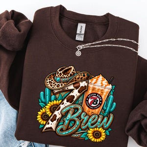 May include: A dark brown crewneck sweatshirt featuring a graphic print of a leopard print cowboy hat, a coffee drink with a Seven Brew logo, sunflowers, and teal cacti. The number seven is patterned with cow print and teal accents.
