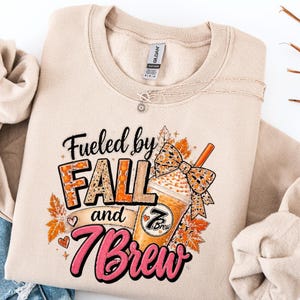 Può includere: Una felpa girocollo beige con una stampa grafica recante il testo "Fueled by FALL and 7 Brew". Il design include una tazza di caffè con un fiocco leopardato, foglie autunnali arancioni e piccoli cuori centrati sul petto.
