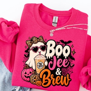 Può includere: Una felpa girocollo rosa acceso con la grafica di un fantasma che indossa occhiali da sole a forma di cuore e un fiocco leopardato, mentre tiene in mano un caffè freddo. Il testo recita Boo Jee & Brew, circondato da zucche e pipistrelli.