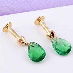 Puede incluir: Un par de pendientes dorados con un poste y una gema verde en forma de lágrima. Los pendientes están colocados sobre un fondo blanco con un toque de morado.