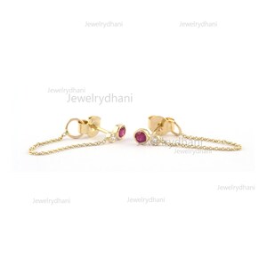 May include: A pair of gold earrings with a small red gemstone and a smaller white gemstone. Each earring has a delicate gold chain dangling from it.