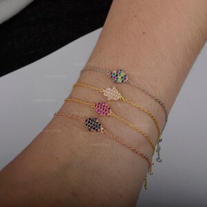 May include: Four gold bracelets with hamsa hand charms. Each charm is encrusted with colorful gemstones. The bracelets are layered on a person's wrist.