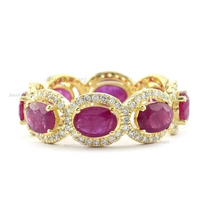 May include: A gold band ring with a row of oval-shaped ruby gemstones set in a halo of smaller diamonds.