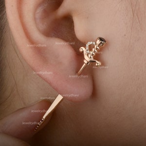 Könnte beinhalten: Ein Paar goldene Ohrringe mit einem Schwertmuster. Ein Ohrring hat einen schwarzen Stein und der andere Ohrring hat eine Reihe kleiner roter Steine.