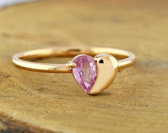 May include: A gold ring featuring a heart-shaped design. The ring has a pink gemstone on one side and a gold heart shape on the other. The ring is set on a wooden surface.