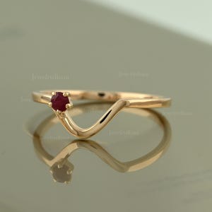 May include: A rose gold ring featuring a single, small, round-cut red ruby gemstone set on one side of a curved, V-shaped band. The metal has a polished, reflective finish, and the design creates a distinct wave-like silhouette on the finger.