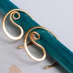 Op de afbeelding: Een paar goudkleurige draad oorbellen met een wervelend ontwerp. De oorbellen worden getoond op een turquoise houten deuvel tegen een zachte, neutrale achtergrond. De oorbellen hebben een minimalistische, elegante uitstraling.