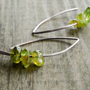 Op de afbeelding: Een paar zilveren oorbellen met groene edelsteen kralen. De oorbellen zijn gemaakt van dunne draad en hebben een eenvoudig, elegant ontwerp.