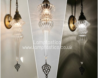 May include: A wall sconce featuring a brass finish mount and a long, ribbed, clear glass shade with a teardrop shape. A decorative brass filigree ornament hangs from the bottom tip. The text Lamptastico www.lamptastico.co.uk is visible.