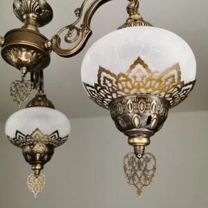 May include: A gold-toned chandelier with three white frosted glass shades. Each shade has a decorative metal design and a heart-shaped pendant.
