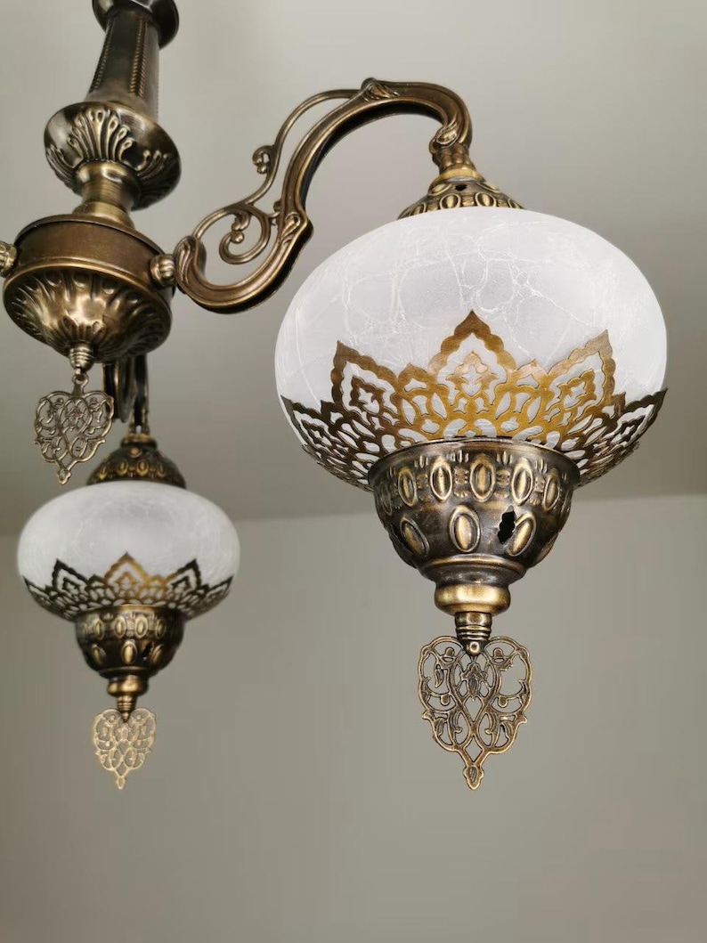 May include: A gold-toned chandelier with three white frosted glass shades. Each shade has a decorative metal design and a heart-shaped pendant.