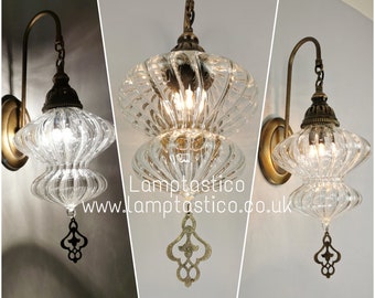 May include: A wall-mounted lamp featuring a clear, ribbed glass shade with a double-bulbous shape. The fixture has an antique brass-toned metal arm and a decorative metal finial hanging from the bottom. Text reads Lamptastico www.lamptastico.co.uk.