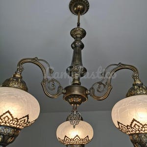 May include: A three-light chandelier with a brass finish and white frosted glass shades. The shades have intricate cut-out designs and are suspended from ornate metal arms.