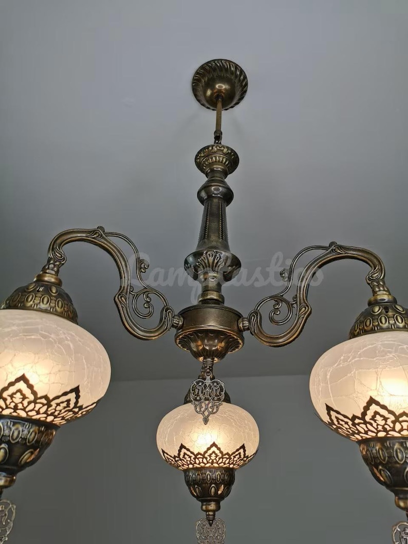 May include: A three-light chandelier with a brass finish and white frosted glass shades. The shades have intricate cut-out designs and are suspended from ornate metal arms.