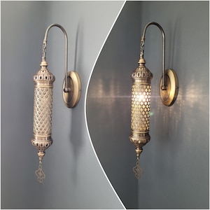 May include: Two brass wall sconces with intricate, geometric designs. Each sconce features a cylindrical shade with a decorative, openwork pattern. The sconces are mounted on a wall with a grey background.