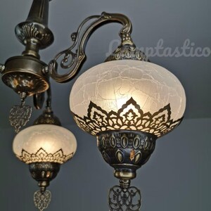 May include: A close-up of a chandelier with three white glass shades. The shades have a decorative, lacy pattern and are illuminated from within. The chandelier is made of brass and has a traditional design.