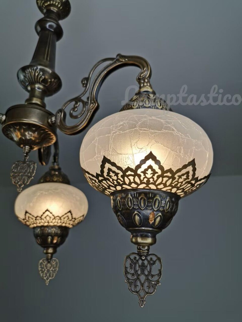May include: A close-up of a chandelier with three white glass shades. The shades have a decorative, lacy pattern and are illuminated from within. The chandelier is made of brass and has a traditional design.