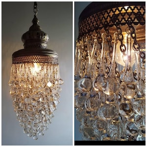 Puede incluir: Una lámpara de araña de latón con un diseño en cascada de cristales de vidrio transparentes en forma de lágrima. La lámpara de araña tiene un diseño tradicional con una parte superior redonda y adornada y un marco de metal.