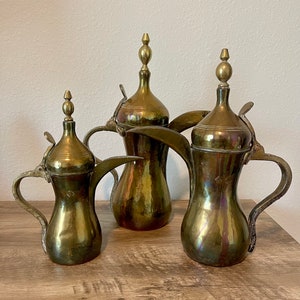 May include: Three brass teapots of varying sizes with ornate spouts and lids. The teapots are arranged on a wooden surface.