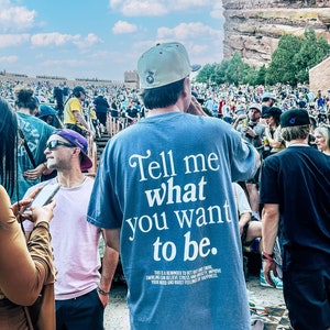 May include: A blue t-shirt with the text "Tell me what you want to be." printed on the front. The text is in a white, cursive font. The shirt also has a small, white text message on the bottom that reads "This is a reminder to get out and swirl. Swirling can relieve stress and anxiety. Improve your mood and boost feelings of happiness."