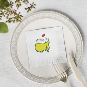 May include: A white paper napkin with the Masters logo, featuring a yellow map of the U.S. with a red flag, is placed on a white plate with a silver rim. A fork and knife are positioned to the right of the napkin. The word "Masters" is written above the map.