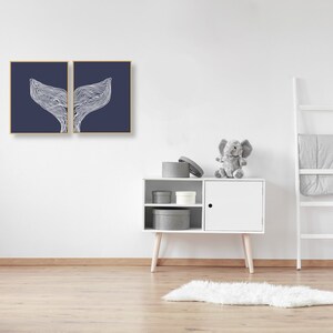 May include: Two framed prints of a white whale tail design on a dark blue background. The prints are hanging on a white wall above a white cabinet with gray storage boxes. A gray stuffed elephant sits on top of the cabinet.