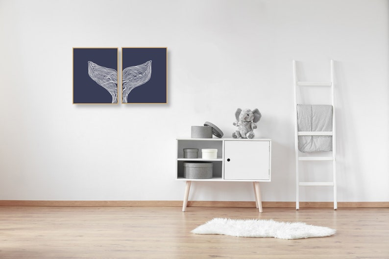 May include: Two framed prints of a white whale tail design on a dark blue background. The prints are hanging on a white wall above a white cabinet with gray storage boxes. A gray stuffed elephant sits on top of the cabinet.
