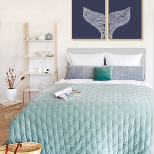 May include: Two framed prints of a white line drawing of a whale tail on a dark blue background. The prints are hanging on a white wall above a bed with a light blue quilted bedspread.
