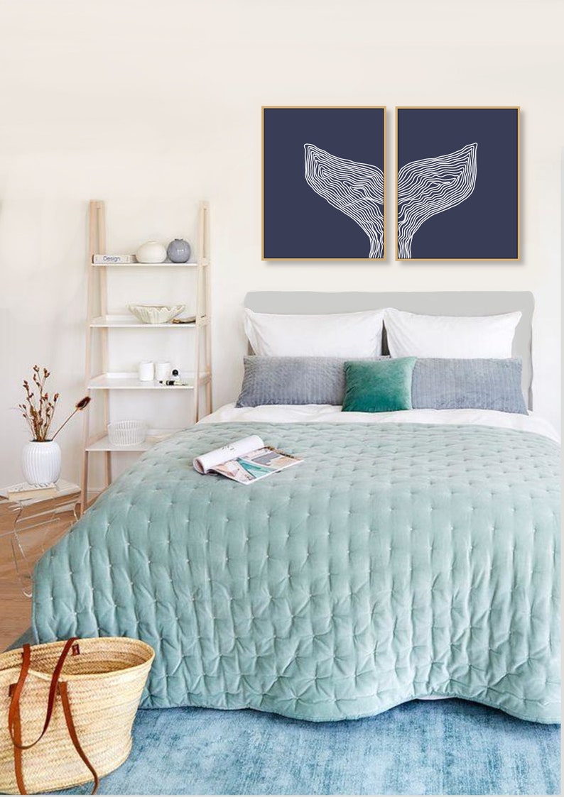 May include: Two framed prints of a white line drawing of a whale tail on a dark blue background. The prints are hanging on a white wall above a bed with a light blue quilted bedspread.