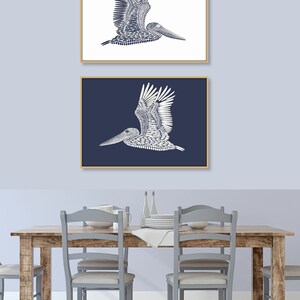 May include: Two framed prints of a white pelican silhouette against a blue background. The top print has a white background and the bottom print has a dark blue background.