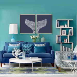 May include: A blue sofa with blue and white patterned pillows sits in front of a teal wall. Two framed prints of white whale tails on a dark blue background hang above the sofa. A white bookshelf with books and decorative items is on the right side of the image. A white coffee table with a white coral sculpture and a blue glass vase sits in front of the sofa. A blue and white patterned armchair sits on the right side of the image.
