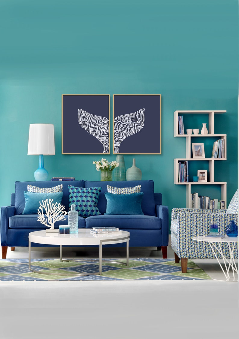 May include: A blue sofa with blue and white patterned pillows sits in front of a teal wall. Two framed prints of white whale tails on a dark blue background hang above the sofa. A white bookshelf with books and decorative items is on the right side of the image. A white coffee table with a white coral sculpture and a blue glass vase sits in front of the sofa. A blue and white patterned armchair sits on the right side of the image.