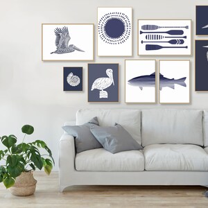 May include: A gallery wall featuring a variety of blue and white prints with nautical themes. The prints include a pelican, a heron, a fish, a seashell, and a sun with oars. The prints are arranged in a symmetrical pattern on a white wall.