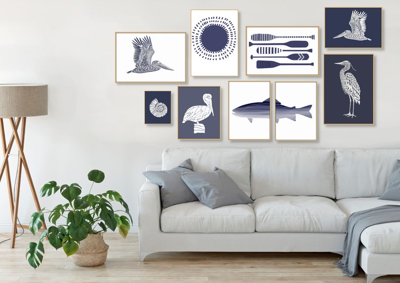 May include: A gallery wall featuring a variety of blue and white prints with nautical themes. The prints include a pelican, a heron, a fish, a seashell, and a sun with oars. The prints are arranged in a symmetrical pattern on a white wall.