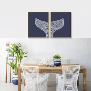 May include: Two framed prints of white whale tail silhouettes on a navy blue background. The prints are hanging on a white brick wall above a wooden table with white wicker chairs.