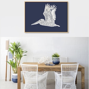 May include: A white pelican silhouette on a dark blue background, framed in wood. The pelican is made up of small white dots. The image is hanging above a wooden table with white wicker chairs.