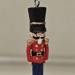 Può includere: Una statuetta di schiaccianoci rossa e dorata con un cappello nero e pantaloni blu pende da un gancio per orecchini d'argento.