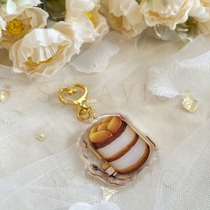 以下が含まれることがあります： 層状のデザートの漫画イラストとスプーンを持った小さなキャラクターが描かれた、透明なアクリルキーホルダー。キーホルダーには、金色のハート型の留め金が付いています。デザートは白、茶色、黄色の層になっています。