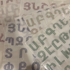 May include: Sheets of clear plastic with various letters made of rhinestones in different colors, including red, blue, green, and purple.