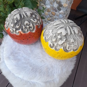 Może przedstawiać: Dwie dekoracyjne ceramiczne kule, jedna czerwona i jedna żółta, z rzeźbionym, teksturowanym wzorem na górnej połowie. Dolne połówki są w jednolitych kolorach. Kule spoczywają na białej, puszystej powierzchni.