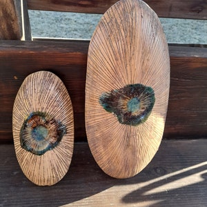 Könnte beinhalten: Zwei handgefertigte, ovale Keramikstücke. Das größere Stück ist etwa 46 cm lang, das kleinere etwa 25 cm. Beide weisen ein radiales Muster aus geätzten Linien und einen zentralen, glasierten Bereich mit Türkis- und Brauntönen auf. Die Stücke liegen auf einer Holzoberfläche.
