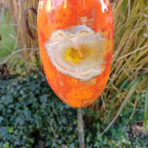 Op de afbeelding: Een levendige, oranje keramische tuinpaal met een uniek, abstract ontwerp. De paal heeft een centraal, oogachtig patroon met gele en witte accenten. De glanzende afwerking en de metalen staaf dragen bij aan de decoratieve uitstraling.
